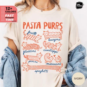 May include: An ivory t-shirt featuring a graphic print with the text PASTA PURRS and illustrations of cats shaped like various pasta types including fusilli, lasagna, fettuccine, cavatappi, penne, macaroni, and spaghetti in orange and blue ink.