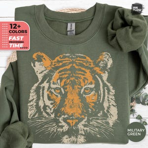 Pode incluir: Uma camisola de gola redonda verde-azeitona com uma grande impressão gráfica central de um rosto de tigre em tons de laranja e creme. A camisola tem mangas compridas, uma gola canelada e uma etiqueta da marca Gildan no interior.