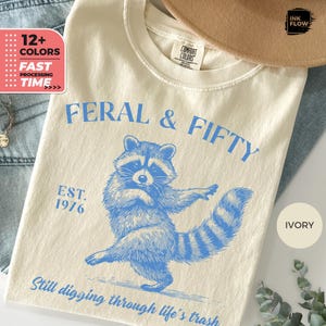 May include: Ivory t-shirt with a blue raccoon graphic and the text "FERAL & FIFTY". The shirt also features the text "EST. 1976" and "Still digging through life's trash". The shirt is made of a soft, comfortable material.