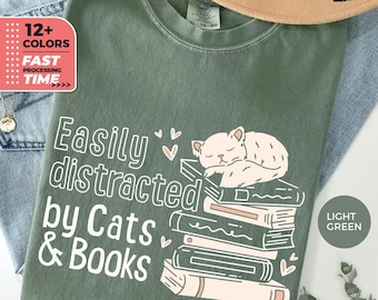 May include: A light green t-shirt with the text "Easily distracted by Cats & Books" in white. A cartoon cat is sleeping on a stack of books.