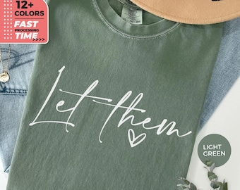 May include: Light green t-shirt with the text "Let them" in white script. The shirt is folded, with a tan hat and blue jeans in the background. A sticker indicates "12+ COLORS" and "FAST PROCESSING TIME."