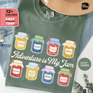 Peut inclure: T-shirt vert clair avec un motif coloré de huit pots de confiture, chacun avec un mot inspirant. La phrase "Adventure is My Jam" est imprimée en dessous. Le t-shirt affiche également le texte "12+ COLORS" et "FAST PROCESSING TIME."