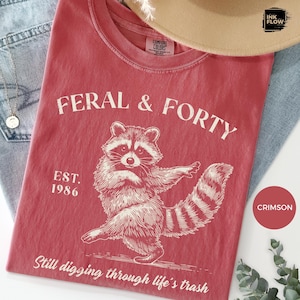May include: Crimson t-shirt with the text "FERAL & FORTY" and a raccoon illustration. The shirt also features the text "EST. 1986" and "Still digging through life's trash". The t-shirt is made of a soft material.