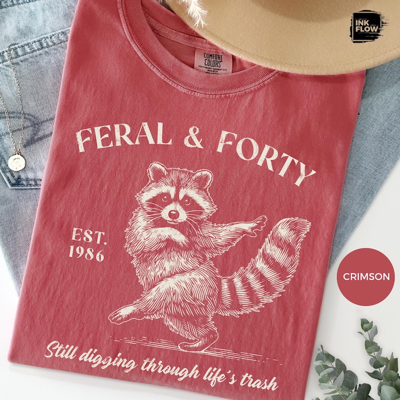 May include: Crimson t-shirt with the text "FERAL & FORTY" and a raccoon illustration. The shirt also features the text "EST. 1986" and "Still digging through life's trash". The t-shirt is made of a soft material.
