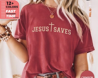 May include: Crimson-colored t-shirt with the text "JESUS SAVES" in a gold color, featuring a cross. The shirt has short sleeves and a crew neck. The image also shows a brown belt and the words "12+ COLORS" and "FAST PROCESSING TIME".