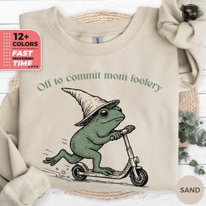 Pode incluir: Camisola bege com um sapo verde usando um chapéu de bruxo e andando de scooter. O texto "Off to commit mom foolery" está acima do sapo. A camisola também tem uma etiqueta "12+ COLORS" e "FAST PROCESSING TIME".