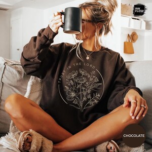 May include: Chocolate-colored sweatshirt with the text "TRUST IN THE LORD" encircling a floral design. The sweatshirt is a solid color and has a crew neck. A black mug is held up to the face.