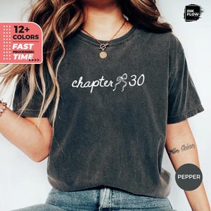 May include: Dark gray t-shirt with the words "chapter 30" in white script. The shirt has a relaxed fit and is made of a soft material. The shirt also has a bow graphic next to the number 30.