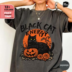 May include: A charcoal grey t-shirt featuring a graphic print of a black cat sitting on a jack-o'-lantern pumpkin in front of a large orange moon. The text "BLACK CAT ENERGY" is printed in a distressed black font above the cat illustration.
