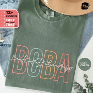 Op de afbeelding: Een lichtgroen T-shirt met de tekst "Board Certified Behavior Analyst" en het acroniem "BCBA" in kleurrijke omlijnde letters. De tekst is in een vintage lettertype.