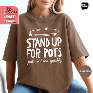Op de afbeelding: Een bruin T-shirt met korte mouwen en witte tekst die STAND UP FOR POTS just not too quickly leest, met een afbeelding van een cirkelvormige bewegingslijn en drie kleine sterren boven de hoofdtekst op het borstgedeelte.