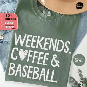 May include: Light green t-shirt with the text "WEEKENDS. COFFEE & BASEBALL." in white. The shirt also has a heart graphic. The shirt is displayed with a hat and jeans. The shirt is available in 12+ colors.