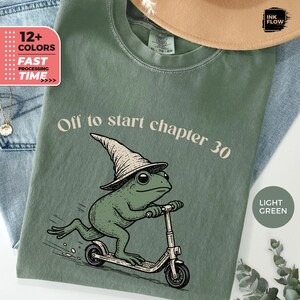 May include: A light green t-shirt featuring a graphic of a frog wearing a pointed hat and riding a scooter. The text "Off to start chapter 30" is printed above the frog. The shirt has a crew neckline and short sleeves, with a visible brand label.