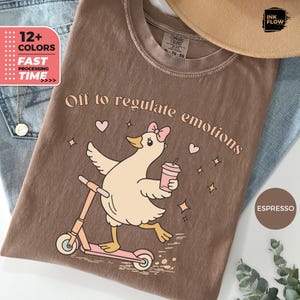 Op de afbeelding: Een espresso-kleurig t-shirt met een afbeelding van een lichtgekleurde eend met een roze strik op een roze step, terwijl deze een roze beker vasthoudt. De tekst "Off to regulate emotions" staat erboven, omringd door kleine hartjes en sterretjes.