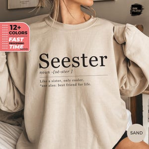 以下が含まれることがあります： 砂色のスウェットシャツ。黒字で「Seester」の文字と定義がプリントされています。「Like a sister, only cooler.」と「see also: best friend for life.」と書かれています。