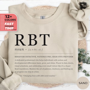 May include: Sand-colored sweatshirt with the letters "RBT" in large, bold, black font. Below the letters is the word "noun" and a definition. The sweatshirt also has text that reads "12+ COLORS" and "FAST PROCESSING TIME".