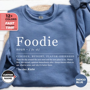 Peut inclure: Sweat-shirt bleu indigo avec le mot "Foodie" en blanc et sa définition. Le texte "Voir aussi : Kate" est également présent. Le sweat-shirt a un col rond et des manches longues, idéal pour un look décontracté.
