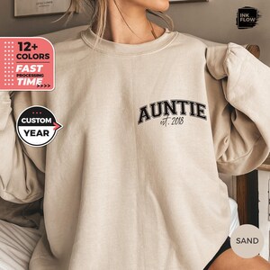 以下が含まれることがあります： 黒い文字で「AUNTIE est. 2018」とプリントされたベージュのスウェットシャツです。