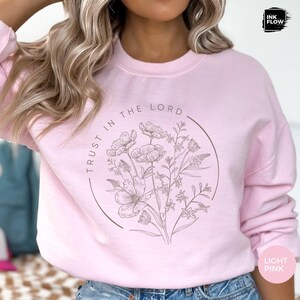 May include: Light pink sweatshirt with a floral design inside a circular frame. The words "TRUST IN THE LORD" are printed around the circle. The sweatshirt also has the words "LIGHT PINK" printed on it.