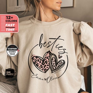 以下が含まれることがあります： ベージュのトレーナーに、黒とピンクのグラフィックデザインが施されています。デザインには「besties」の文字と、2つのハートが描かれており、1つのハートにはレオパード柄が施されています。円の中に「Since 2019」の文字があり、ハートの下には「Emma Olivia」の名前が書かれています。