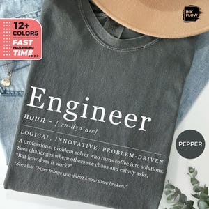 May include: A dark grey t-shirt featuring the definition of "Engineer" in white text, including the noun, pronunciation, and a description. The shirt is shown with a tan hat and blue jeans. The shirt also has the text "12+ COLORS" and "FAST PROCESSING TIME".