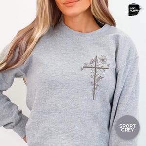 May include: A grey sweatshirt featuring a brown floral cross design on the left chest. The flowers and cross are in shades of brown. The sweatshirt is labelled "Sport Grey" in a circle on the lower right.