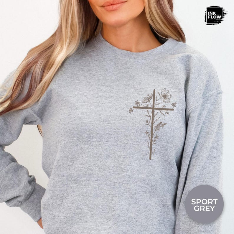 May include: A grey sweatshirt featuring a brown floral cross design on the left chest. The flowers and cross are in shades of brown. The sweatshirt is labelled "Sport Grey" in a circle on the lower right.