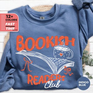 May include: Indigo blue sweatshirt with the words "BOOKISH READERS CLUB" in orange and white. The design includes an open book, a coffee cup with a heart, and decorative bows. The sweatshirt has a crew neck.