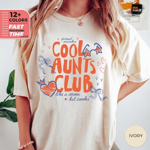 May include: Ivory-colored t-shirt with the text "Cool Aunts Club" in orange, blue, and red. The design includes stars, hearts, and bows. The shirt also has the text "like a mom but cooler".