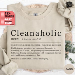 May include: A sand-colored crewneck sweatshirt featuring the black printed text Cleanaholic, defined as a noun with phonetic spelling, followed by a descriptive paragraph about being organized, detail-obsessed, and cleaning-powered.