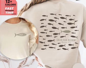 May include: Sand-colored sweatshirt with a fish graphic on the front and back. The back features a school of fish with one fish in a different color, and the text "CALLED TO BE DIFFERENT Romans 12:2". The front has a fish graphic and the text "ROMANS 12".