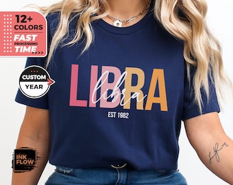 May include: A navy blue t-shirt with the word "LIBRA" in pink, orange, and yellow letters. The word "libra" is written in white script underneath the letters. The text "EST 1982" is printed below the word "libra".