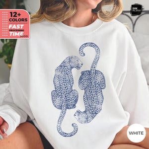 May include: A white crewneck sweatshirt featuring a dark blue graphic print of two leopards positioned vertically. The leopards are depicted with detailed spots and long tails, one facing right and the other facing left, centered on the chest.