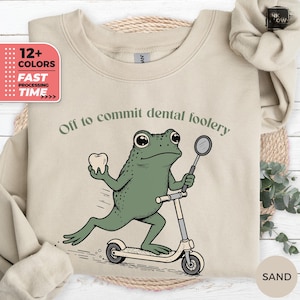 May include: A sand-coloured crewneck sweatshirt featuring a graphic of a green frog riding a kick scooter while holding a white tooth. The text above the frog reads, "Off to commit dental foolery." The design is printed in dark green and black ink.