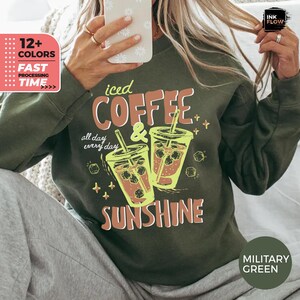 Könnte beinhalten: Militärgrünes Sweatshirt mit dem Schriftzug "iced COFFEE & SUNSHINE" in einem Retro-Schriftstil. Die Grafik zeigt zwei Eiskaffee-Getränke mit Strohhalmen. Das Sweatshirt hat auch den Text "all day every day".