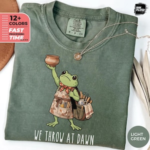 May include: A sage green t-shirt featuring a graphic of a frog wearing an apron and carrying a bag of pottery tools. The frog holds a clay bowl above its head. Below the graphic, the text reads WE THROW AT DAWN. A gold chain necklace rests on the collar.