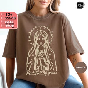 May include: Espresso-colored t-shirt featuring a cream-colored graphic of a religious figure with a halo and the text "Hail! full of grace." The shirt has short sleeves and a relaxed fit. The image also includes text indicating multiple color options and fast processing time.
