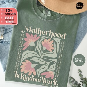 May include: Light green t-shirt featuring the text "Motherhood Is Kindom Work" and a floral design in pink and light green. The shirt also has the text "PROV. 31:25-24". The shirt is displayed with a hat and jeans.