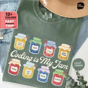 May include: Light green t-shirt with the phrase "Coding is My Jam" and illustrations of jam jars. Each jar contains a different fruit and a coding-related word like "Debug" or "Logic". The shirt also has a tag that says "12+ Colors".