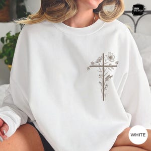 May include: White sweatshirt with a brown cross design decorated with floral and butterfly details. The sweatshirt has a crew neck and long sleeves. The word "WHITE" is printed on the bottom right corner.
