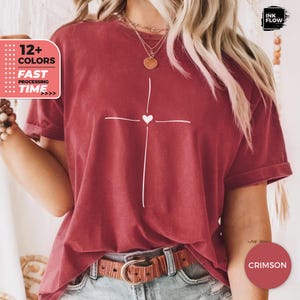 May include: A crimson short-sleeved t-shirt featuring a white line-art cross graphic with a small heart at the centre. The shirt is worn with a brown leather belt and a layered gold necklace. Text on the image includes 12+ COLORS, FAST PROCESSING TIME, INK FLOW, and CRIMSON.