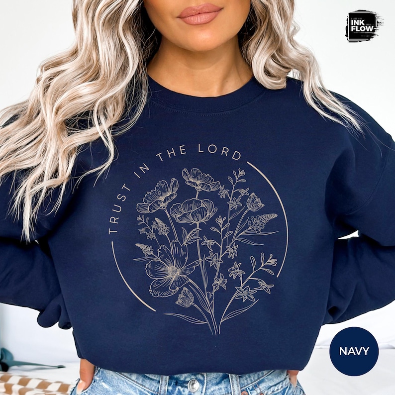 May include: Navy blue sweatshirt featuring a floral design within a circular frame. The text "TRUST IN THE LORD" is arched around the floral illustration. The word "NAVY" is in a white box in the lower right corner.