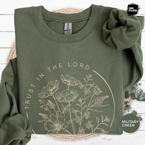 May include: Military green sweatshirt with the text "TRUST IN THE LORD" and a floral design in a circular shape. The sweatshirt is made of a soft material and has a crew neck. The Ink Flow logo is in the top right corner.