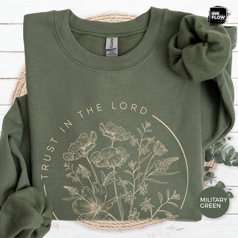 May include: Military green sweatshirt with the text "TRUST IN THE LORD" and a floral design in a circular shape. The sweatshirt is made of a soft material and has a crew neck. The Ink Flow logo is in the top right corner.