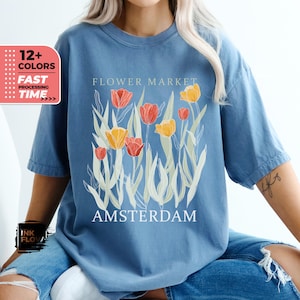 Może przedstawiać: Niebieska koszulka z grafiką stylizowanych tulipanów i tekstem "FLOWER MARKET AMSTERDAM".