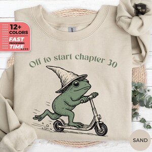 May include: A sand-colored crewneck sweatshirt featuring a graphic of a green frog wearing a pointed hat and riding a kick scooter. The text above the frog reads, "Off to start chapter 30." The sweatshirt has long sleeves and a ribbed neckline.