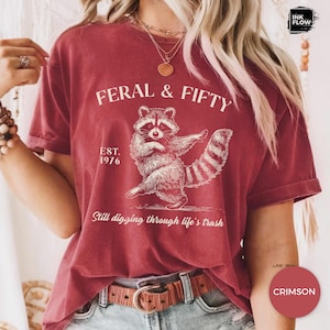 May include: Crimson-colored t-shirt with a raccoon graphic and the text "FERAL & FIFTY". The raccoon illustration is in white, with the text "EST. 1976" and "Still digging through life's trash" below the raccoon.