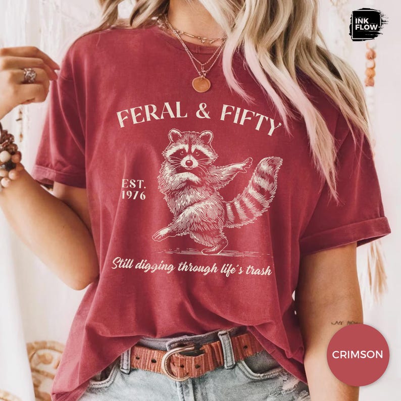 May include: Crimson-colored t-shirt with a raccoon graphic and the text "FERAL & FIFTY". The raccoon illustration is in white, with the text "EST. 1976" and "Still digging through life's trash" below the raccoon.