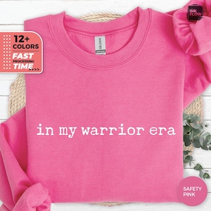 May include: A pink sweatshirt with the text "in my warrior era" printed in white on the front.