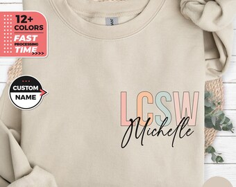 Könnte beinhalten: Ein beiger Pullover mit dem Text "LCSW Michelle" in bunten Buchstaben. Die Buchstaben sind schwarz umrandet und haben ein pastellfarbenes Farbschema. Der Pullover liegt auf einer hellbraunen Oberfläche.
